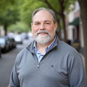 The author, Eric L Dickey, a white male with short greying hair combed back, sporting a neatly trimmed short grey beard with mustache, wearing a blue gingham business casual dress shirt under a gray quarter-zip sweater on a tree-lined city street.