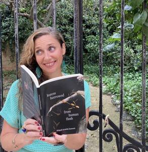 A woman looking sideways and holding her latest book 'Leaves Borrowed from Human Flesh', with background leaves and gate.