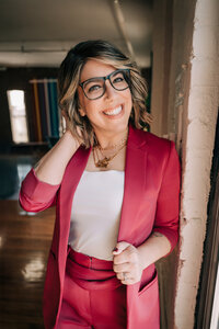 photograph of Kathryn Nolan, a white person with short, wavy brunette hair, glasses, and a pink blazer on with a white tank top underneath.
