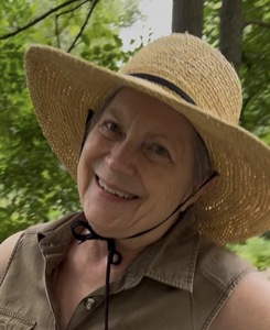 Headshot of the smiling author wearing a floppy, tan hat and a safari-type sleeveless blouse. Woods & trees in background.