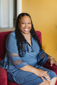 Afrika sitting in a reddish chair with a bluish dress on smiling