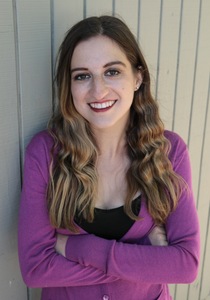 a woman with dark hair and purple sweater leans against a building