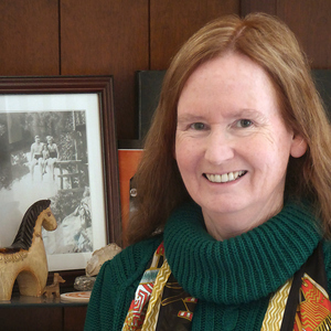 A female-presenting person with light skin and long reddish-brown hair, wearing a colorful scarf and a dark green sweater smiles into the camera. In the background, a figure of a horse and a black-and-white photo of a family group.