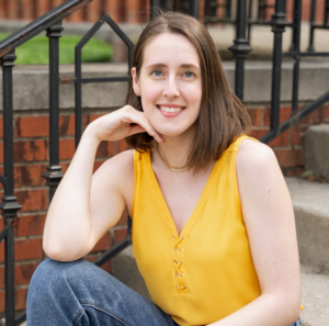 Shelby smiling in a bright yellow blouse and jeans. Her hair is shoulder length and brown, and her hand is below her chin.