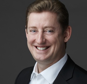Darren B. Todd, smiling, in a black jacket and open-collar white shirt against a dark gray studio background.