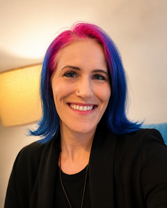 Kat Kennan smiling at the camera, wearing a black blazer with bright pink and blue hair.