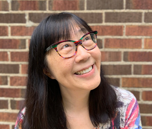 A woman with long dark hair and colorful, patterned glasses smiles while looking up, standing in front of a brick wall. She is wearing a colorful top that complements the vibrant tones of her glasses. The expression on her face appears joyful and thoughtf