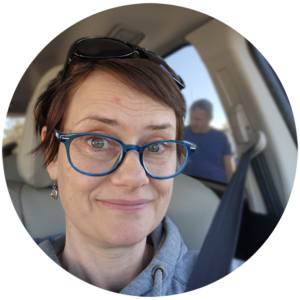 Author Jenn Shon headshot selfie from the passenger seat with a blurred out figure outside over her shoulder