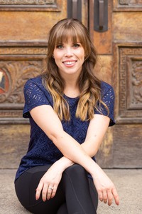 alt="Professional headshot of a smiling woman with long, wavy brown hair and bangs, wearing a navy lace top, seated in front of a detailed wooden door."