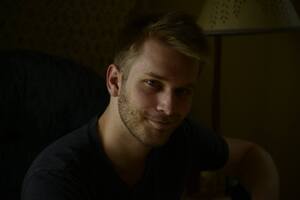  Young man with short blond hair and a trimmed beard, wearing a dark shirt, sitting indoors in low lighting with a soft smile and a relaxed posture.