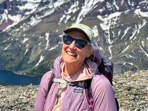 Becky is in hiking attire with a pack in front of a mountain with lingering snow. 