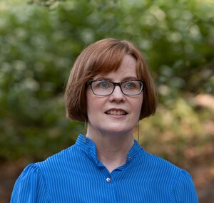 Photo of Kate Deimling, a White woman with short red hair and a blue shirt, in front of trees in a park.