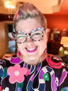 Harlan DeChamps - a female-presenting person in a floral dress, washed out color in their pulled up hair, and checkered glasses, gives a goofy and skewed grin to the camera