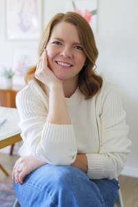 Mira larkin sits in a chair leaning forward with her hand resting on the side of her face. She smiles warmly into the camera.
