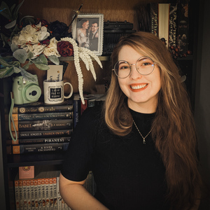 Moody photo of author Xanna Renae standing in front of a packed bookshelf. Xanna has long reddish hair, round green glasses, and is wearing a black short sleeve high neck shirt. 