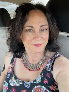 Photo of a smiling white woman in her 50s wearing a multicolor necklace and black tank top with sugar skulls, sitting in the driver's seat of a car