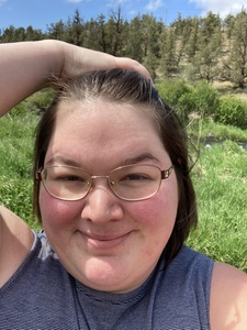 A woman with short brown hair and wire glasses smiling while standing outside. Her hand in on top of her head, holding hair away from her face.