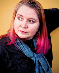 Closeup portrait of a white woman in her 40s. She has shoulder-length brown hair that has been dyed pink at the ends. She is wearing a gray sweater and a lighter gray scarf. Behind her is a yellow wall. She is looking offscreen.