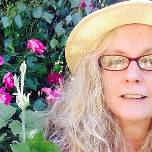 A close-up of woman wearing a hat and glasses in front of a rosebush.