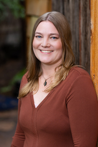 A woman with shoulder length blonde hair wearing a brown shirt, with pine trees in the background. 