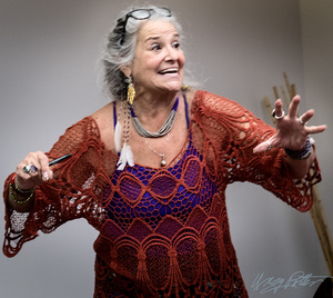 Judyth Hill, poet and writing teacher, animatedly sharing a story with an expressive gesture, wearing a rust-red crocheted shawl and purple top.