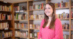 Woman in pink shirt with long brown hair smiles against a background of books.