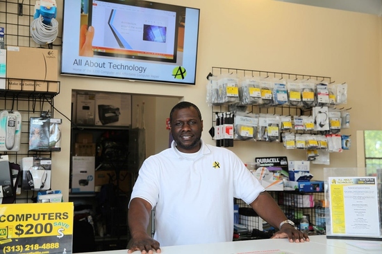 Willie Brake stands smiling behind the front counter of All About Technology in Detroit, surrounded by cables, accessories, and a "Computers $200" sign