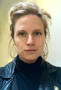 an image of a blonde woman with her hair in a bun and blue eyes, wearing a leather jacket, hoop earrings and a light smile