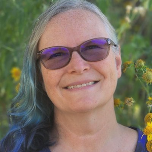 Paige E. Ewing smiling with dark glasses and greenery and yellow daisies in the background