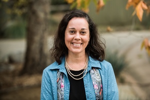 Smiling middle aged white woman with shoulder length brown hair wearing a blue top, multi colored scarf and a strand of pearls. Background is set in a public park surrounded by trees. 