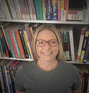 head shot of white woman with blonde hair and red glasses wearing a grey striped shirt standing in front of a bookshelf