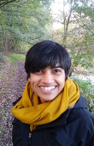 The author is an Indian woman, shown in this photo with short cropped black hair. She had brown eyes. She is smiling at the camera, wearing a mustard yellow scarf and black jacket, on a woodland path.