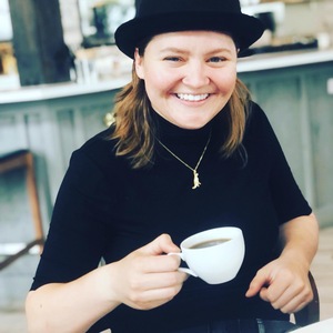 Photo of a woman drinking coffee. She's wearing a bowler hat and a black turtleneck