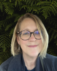 Woman with short blonde hair and large brown glasses, against a background of ferns.