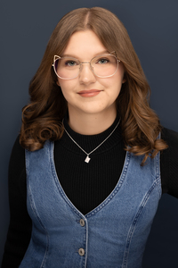 Author Paige N. Regan in a black sweater and blue denim vest standing in front of a blue background