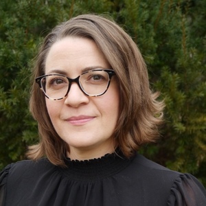 Profile photo of Alia Tawil: Smiling woman with shoulder-length brown hair and glasses, wearing a black, high-necked blouse, standing outdoors  with tall shrubbery in the background.
