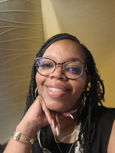 A person with glasses and braided hair smiles at the camera, resting their chin on their hand. They are wearing a black top and gold jewelry.