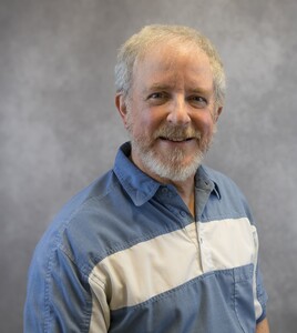 Studio headshot of White male, gray hair and beard, in blue polo shirt with white band across chest.