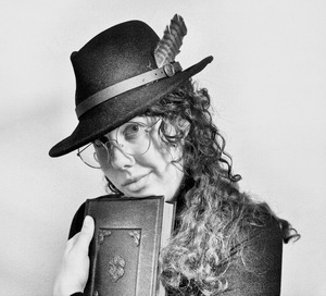 A picture of a white woman with dark curly hair wearing a hat and holding a notebook