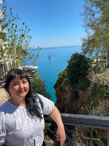 Author standing in front of Katyn Bridge in Antalya, Turkiye. Black long hair, white blouse, behind her is the chasm where the Mediterreanean sea can be seen. 