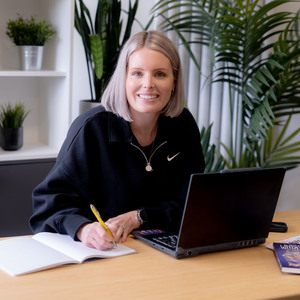 kirstie watson - short blond blob - sitting at a desk - smiling with pen in hand, a notebook and an laptop open.