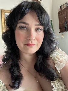 Portrait of a person with dark wavy hair, soft makeup, and a lace dress in natural light.