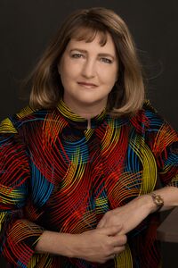 Author in a colorful dress, smiling slightly, leaning on a desk against a dark background.