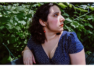 white latina woman with short dark hair, dark eyes, in a polka dot blue dress, looking to the side, against a backdrop of greenery.