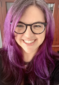 the photo is of a woman with purple hair and glasses. Behind her is a bookcase. 
