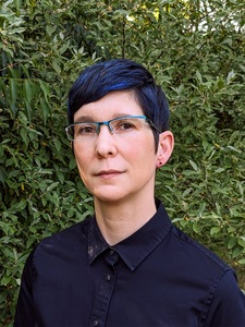 a photo of a light-skinned person with short black hair & blue highlights standing in front of greenery