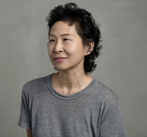 A headshot of an Asian woman with dark, medium-length layered hair and a slight smile wearing a gray tee-shirt.