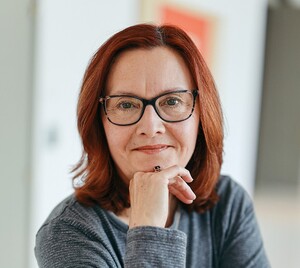 Woman with red hair and glasses, resting her chin on her hand and looking into the camera 