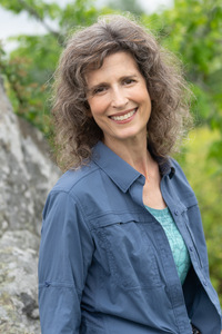 A headshot of a woman with brown wavy hair gazing at the camera, wearing a blue t-shirt & blue long-sleeve shirt over it. She is smiling. She's sitting on a rock with foliage around and behind her.