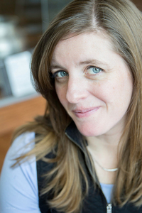 A close-up of a white woman with long brown hair and blue eyes smiles demurely into the camera